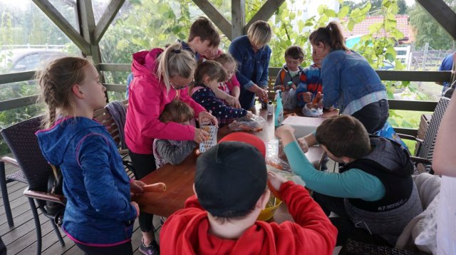 Fotografia przedstawia grupę która znajduje się w altance, gdzie trze na tarce marchewki dla zwierzątek. Dzieciom pomagają rodzice.